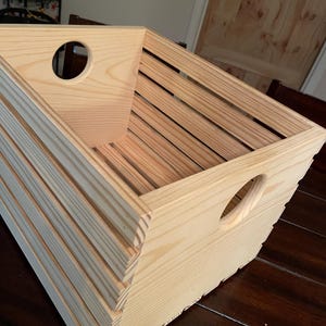 May include: A light-colored wooden storage crate featuring horizontal slats and circular cut-out handles on two opposing sides. The crate has an open top and a rectangular base, constructed from natural wood with visible grain patterns throughout.