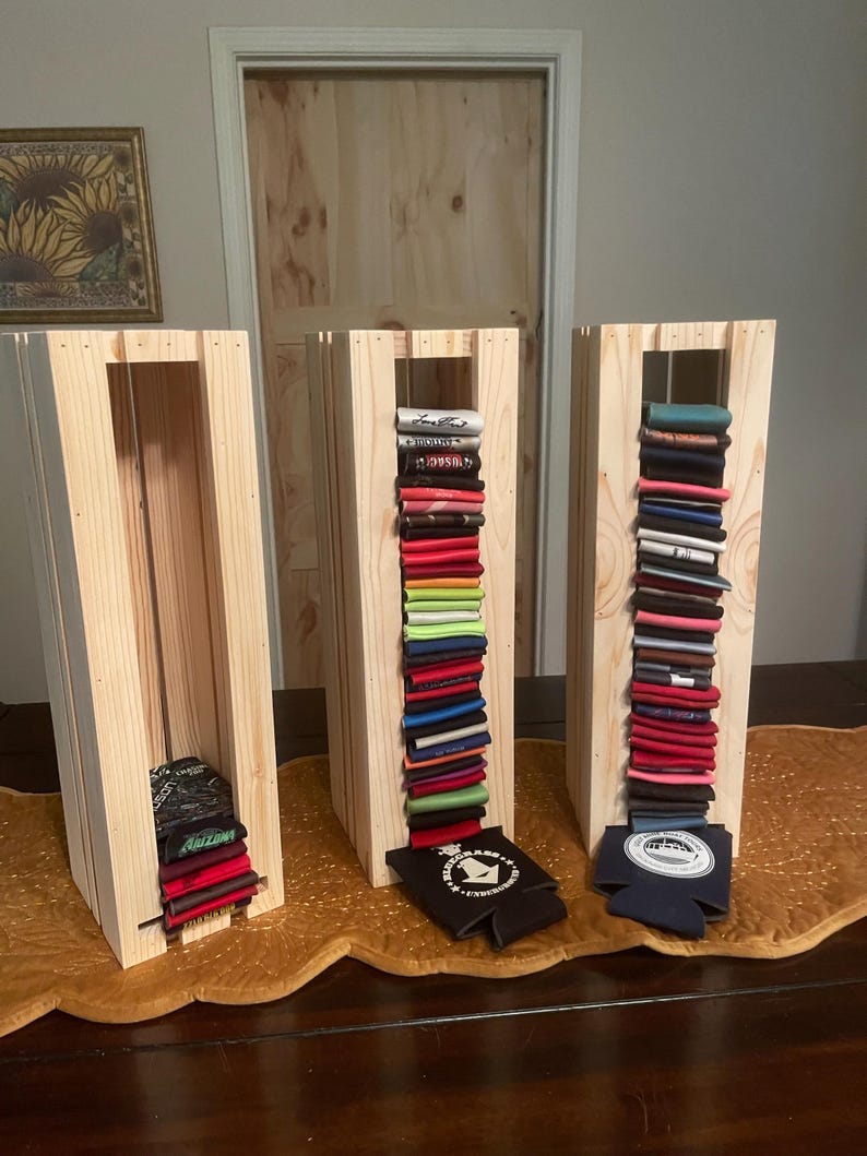 May include: Three wooden display stands filled with colorful beverage can coolers. The stands are made of light-colored wood and are rectangular with vertical slats. The coolers are stacked inside, showcasing a variety of colors and designs.