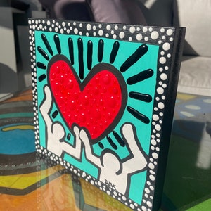 K&ouml;nnte beinhalten: Eine quadratische Leinwand in T&uuml;rkisblau mit schwarzem Rand und wei&szlig;en Punkten. Zwei wei&szlig;e Figuren mit erhobenen Armen halten ein rotes Herz mit wei&szlig;en Punkten. Das Herz ist von schwarzen Strahlen umgeben. Die Leinwand ist unten rechts mit "DDK" signiert.