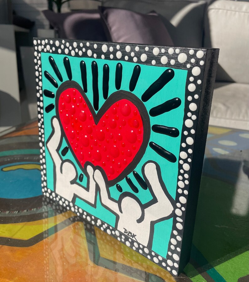 K&ouml;nnte beinhalten: Eine quadratische Leinwand in T&uuml;rkisblau mit schwarzem Rand und wei&szlig;en Punkten. Zwei wei&szlig;e Figuren mit erhobenen Armen halten ein rotes Herz mit wei&szlig;en Punkten. Das Herz ist von schwarzen Strahlen umgeben. Die Leinwand ist unten rechts mit "DDK" signiert.