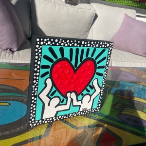 K&ouml;nnte beinhalten: Ein kleines quadratisches Holzst&uuml;ck mit schwarzem Rand und wei&szlig;en Punkten. Die Mitte des Quadrats zeigt einen t&uuml;rkisfarbenen Hintergrund mit einem roten Herzen mit wei&szlig;en Umrissen und einem schwarzen Sonnenstrahl. Zwei wei&szlig;e Figuren mit schwarzen Umrissen halten das Herz hoch. Die Unterschrift "DKK" befindet sich in Schwarz unten rechts.