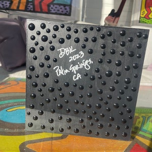 K&ouml;nnte beinhalten: Schwarzes Quadrat mit einer strukturierten Oberfl&auml;che aus erhabenen schwarzen Kreisen. Das Quadrat ist in wei&szlig;er Tinte mit dem Text "DBK 2023 Palm Springs, CA" signiert.