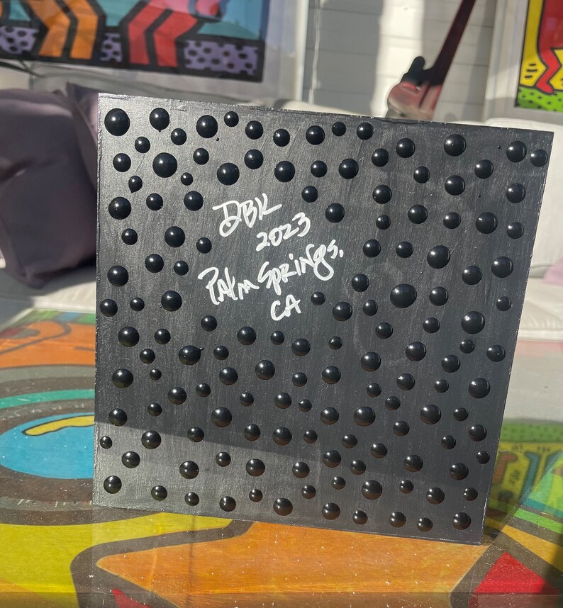 K&ouml;nnte beinhalten: Schwarzes Quadrat mit einer strukturierten Oberfl&auml;che aus erhabenen schwarzen Kreisen. Das Quadrat ist in wei&szlig;er Tinte mit dem Text "DBK 2023 Palm Springs, CA" signiert.