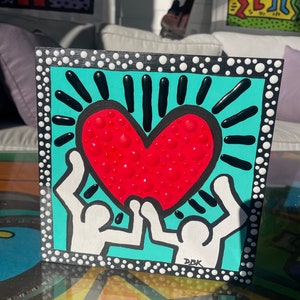 K&ouml;nnte beinhalten: Ein schwarz gerahmtes Gem&auml;lde mit t&uuml;rkisfarbenem Hintergrund. Zwei wei&szlig;e Figuren halten ein rotes Herz mit wei&szlig;en Punkten hoch. Das Herz ist von schwarzen Strahlen umgeben. Das Gem&auml;lde ist unten rechts mit "DBK" signiert.