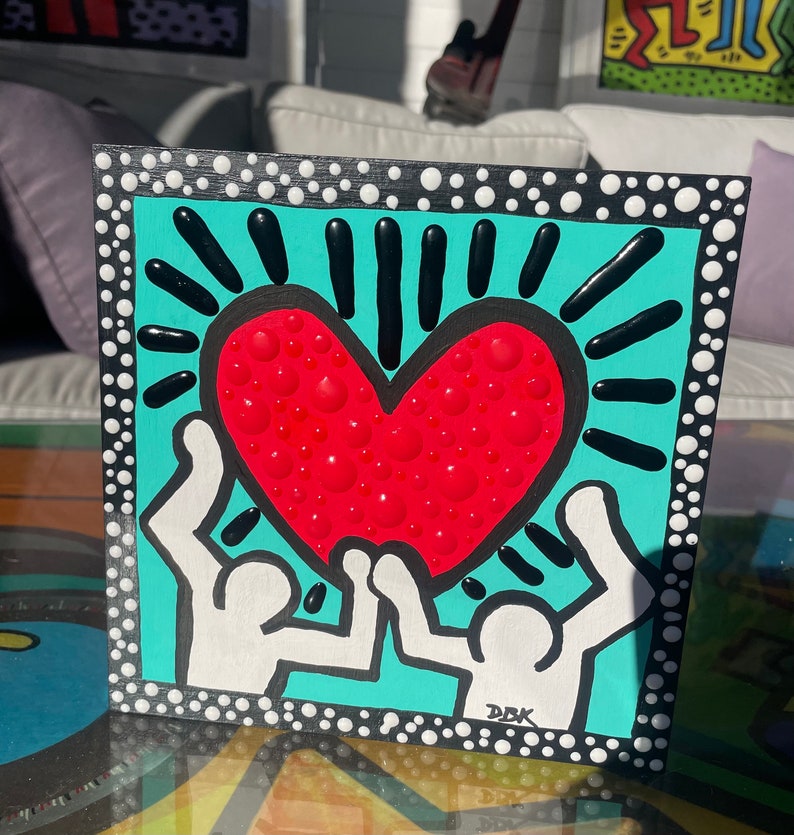 K&ouml;nnte beinhalten: Ein schwarz gerahmtes Gem&auml;lde mit t&uuml;rkisfarbenem Hintergrund. Zwei wei&szlig;e Figuren halten ein rotes Herz mit wei&szlig;en Punkten hoch. Das Herz ist von schwarzen Strahlen umgeben. Das Gem&auml;lde ist unten rechts mit "DBK" signiert.