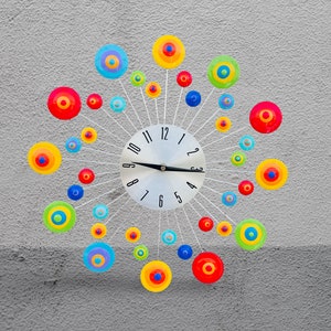 May include: A colorful clock with a white face and black hands. The clock has a unique design with colorful circles around the face. The numbers on the clock face are black.