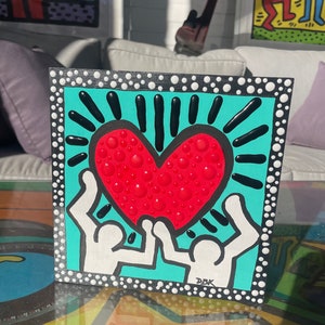 K&ouml;nnte beinhalten: Ein quadratisches Leinwandbild in T&uuml;rkisblau mit einem wei&szlig;en Rand aus wei&szlig;en Punkten. Zwei wei&szlig;e Figuren mit erhobenen Armen halten ein rotes Herz mit wei&szlig;en Punkten. Das Herz ist von schwarzen Linien umgeben, die nach au&szlig;en strahlen. Das Bild ist unten rechts mit "DBK" signiert.