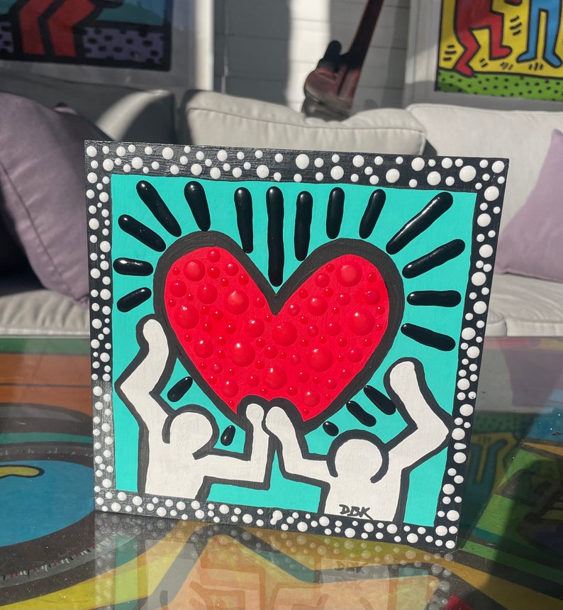 K&ouml;nnte beinhalten: Ein quadratisches Leinwandbild in T&uuml;rkisblau mit einem wei&szlig;en Rand aus wei&szlig;en Punkten. Zwei wei&szlig;e Figuren mit erhobenen Armen halten ein rotes Herz mit wei&szlig;en Punkten. Das Herz ist von schwarzen Linien umgeben, die nach au&szlig;en strahlen. Das Bild ist unten rechts mit "DBK" signiert.