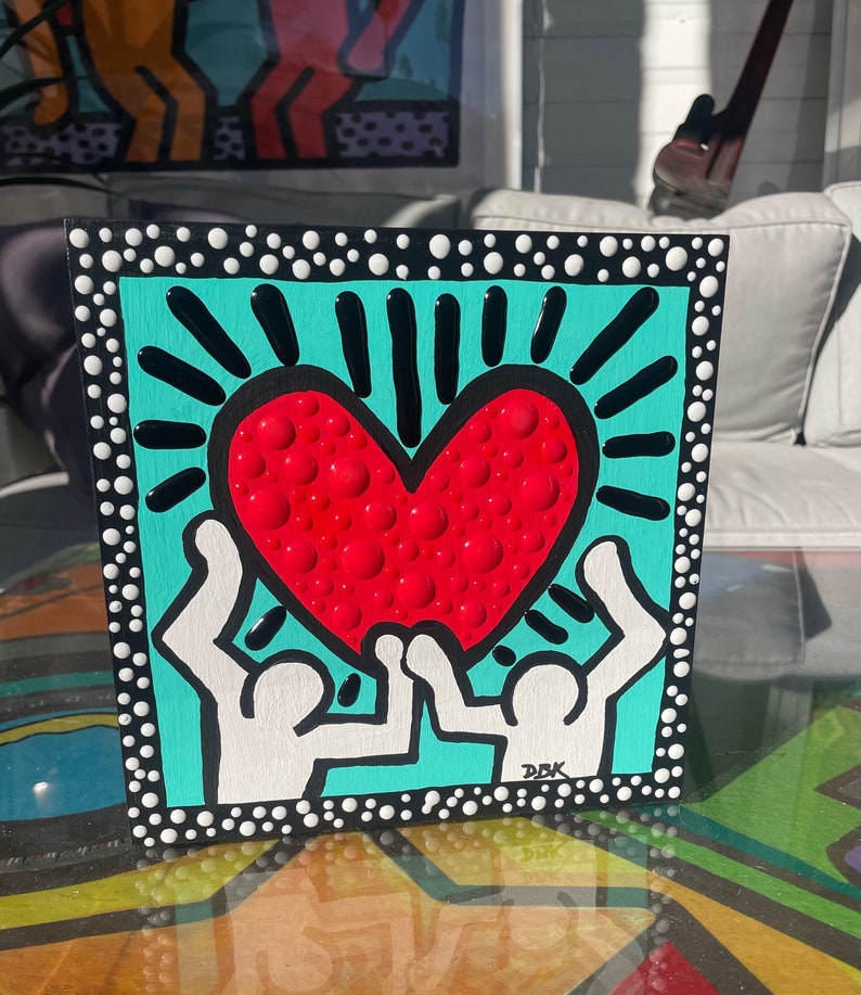 K&ouml;nnte beinhalten: Eine handbemalte Leinwand aus Holz mit t&uuml;rkisfarbenem Hintergrund und wei&szlig;en Punkten. Zwei wei&szlig;e Figuren halten ein gro&szlig;es rotes Herz mit wei&szlig;en Punkten hoch. Das Herz hat schwarze Strahlen, die von ihm ausgehen. Die Leinwand ist unten rechts mit "DBK" signiert.