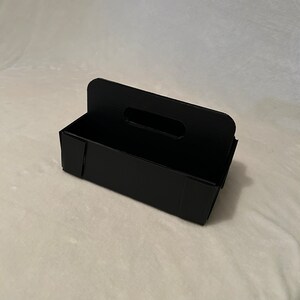 May include: Black cardboard storage box with a handle. The box has a rectangular shape and is made of sturdy cardboard.