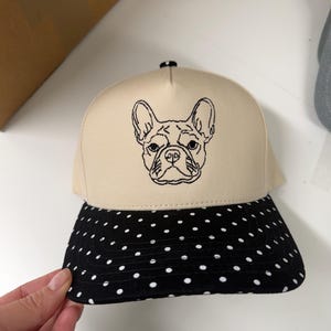 May include: A beige baseball cap with a black and white polka dot peak. The cap has a black embroidered outline of a French Bulldog's face on the front.