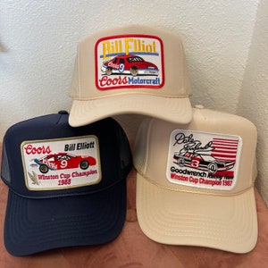 Op de afbeelding: Drie beige en donkerblauwe baseballpetten met geborduurde patches met NASCAR-coureurs Bill Elliott en Dale Earnhardt. De patches tonen de namen van de coureurs, de auto nummers, sponsors en het jaar waarin ze de Winston Cup Championship wonnen.