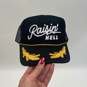 Puede incluir: Gorra de camionero negra con un logotipo bordado en blanco que dice "Raisin' Hell". La gorra tiene detalles bordados en dorado en la visera.