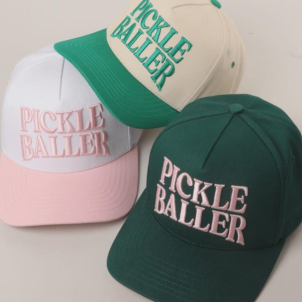 May include: Three baseball caps with the text "PICKLE BALLER" embroidered on the front. One cap is white with a pink brim and pink text, another is cream with a green brim and green text, and the third is dark green with pink text.