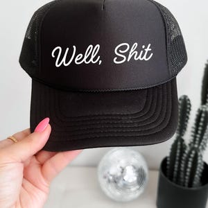 May include: Black trucker hat with a curved brim and mesh back. The hat features the white cursive text "Well, Shit" across the front. The hat is being held by a person with pink nail polish.
