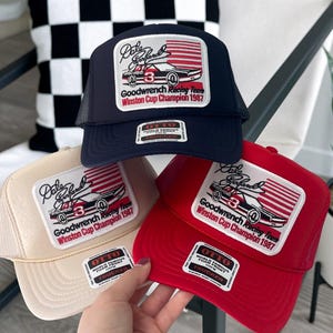 Pode incluir: Três bonés de caminhoneiro com um design de bandeira americana vermelha, branca e azul. Os bonés apresentam uma imagem em preto e branco de um carro de corrida com o texto "Dale Earnhardt" e "Goodwrench Racing Team Winston Cup Champion 1987". Os bonés são de cor azul marinho, bege e vermelho.
