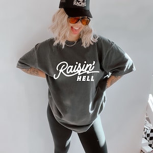 May include: A woman wearing a black t-shirt with the text "Raisin' Hell" in white. She is also wearing black leggings and a black trucker hat with a skull and crossbones logo that says "Dead Last Racing".