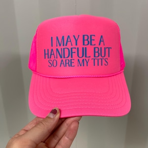 Puede incluir: Gorra de camionero rosa brillante con el texto azul "I MAY BE A HANDFUL BUT SO ARE MY TITS". La gorra tiene una parte trasera de malla y una visera curva.