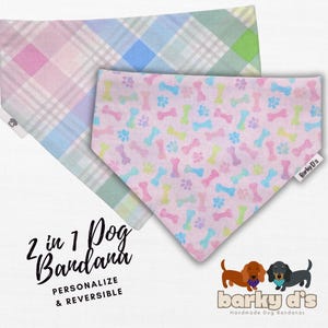 May include: A pink and blue plaid dog bandana with a reversible side featuring a pink background with white paw prints and bone designs. The bandana is labeled "2 in 1 Dog Bandana, Personalize & Reversible" and "Barky D's Handmade Dog Bandanas".