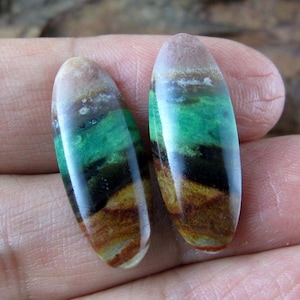 May include: Two polished, oval-shaped gemstones with a layered appearance. The stones feature bands of color, including brown, black, green, and a translucent top. The gemstones are held in a hand, showcasing their natural beauty and unique patterns.