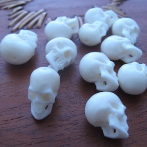 May include: A collection of white skull beads, carved from bone, with intricate details. The beads are smooth and polished, and they are arranged in a random pattern on a wooden surface.