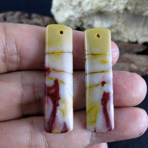May include: Two rectangular gemstone pendants. The pendants are yellow at the top, transitioning to white with red and yellow veining. Each has a small hole at the top for stringing. The stones are held in a hand.