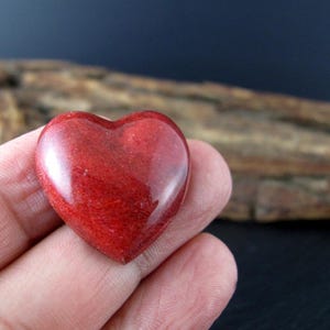 May include: A polished, heart-shaped stone in a deep red hue. The stone has a smooth, glossy surface and is held in a person's hand. The background features a blurred wooden texture and a dark blue backdrop.
