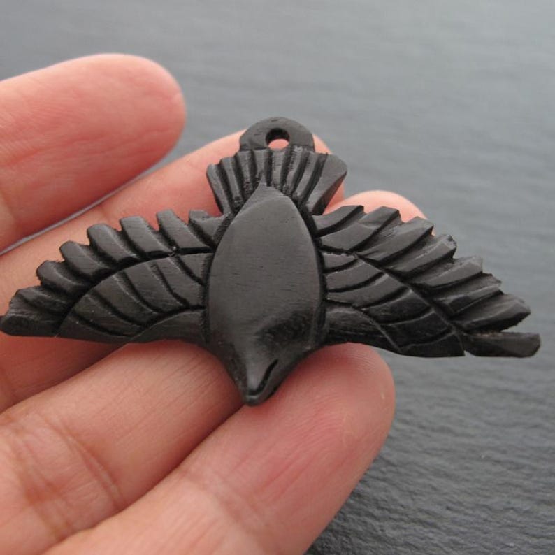 Op de afbeelding: Een zwarte, gesneden houten hanger in de vorm van een vogel met uitgespreide vleugels. De hanger heeft een klein gaatje aan de bovenkant om op te hangen. De vleugels en het lichaam zijn gedetailleerd met gesneden lijnen, wat het een textuur geeft. De vogel is donker van kleur.