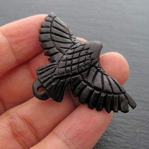 Op de afbeelding: Een gebeeldhouwde zwarte houten adelaar hanger met uitgestrekte vleugels. De adelaar heeft gedetailleerde gebeeldhouwde veren en een kleine lus om op te hangen. De hanger wordt in een hand gehouden, wat het ingewikkelde vakmanschap en ontwerp benadrukt.