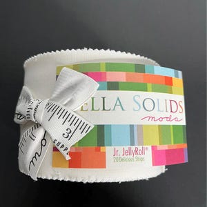 May include: A roll of white fabric strips tied with a white ribbon printed with a ruler design. The label reads "BELLA SOLIDS moda" and "Jr. JellyRoll® 20 Delicious Strips". The ribbon measures 3 inches.