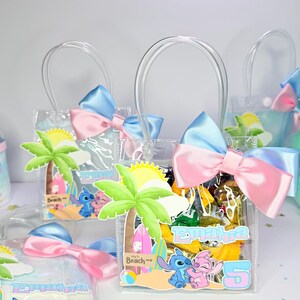 May include: Clear plastic gift bags with blue and pink bows. Each bag features a palm tree, sun, and cartoon characters. The bags have the name "Emahra" and the number "5" printed on them. The bags are filled with treats.