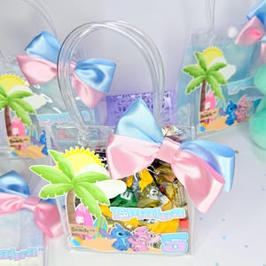 May include: Clear plastic favor bags with blue and pink bows, filled with wrapped candies. Each bag features a beach-themed graphic with a palm tree, sun, and cartoon characters. The bags have clear handles.
