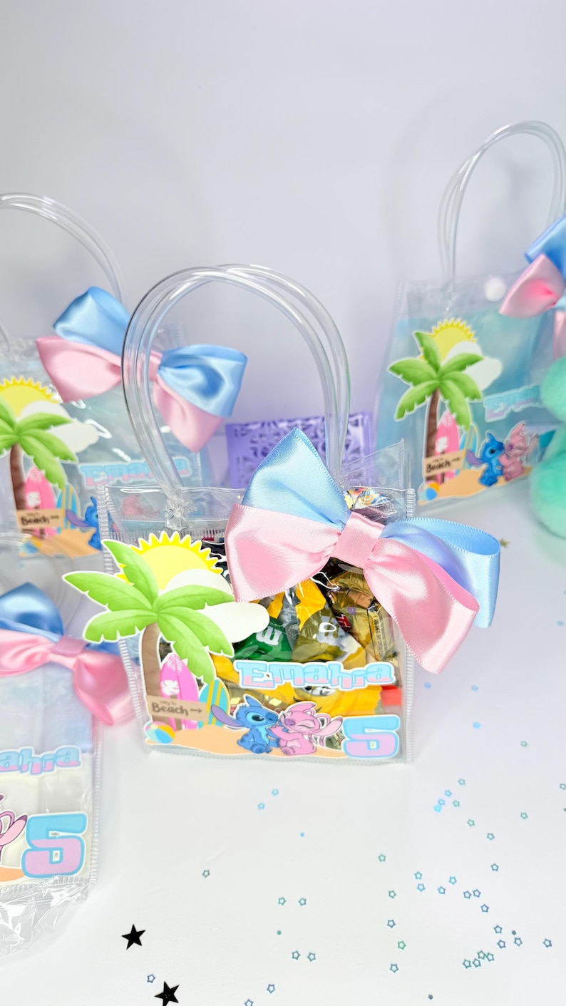 May include: Clear plastic favor bags with blue and pink bows, filled with wrapped candies. Each bag features a beach-themed graphic with a palm tree, sun, and cartoon characters. The bags have clear handles.