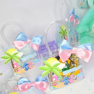 May include: Clear plastic tote bags with blue and pink satin bows. Each bag features a beach-themed graphic with cartoon characters, a palm tree, and the name "Emalra" with the number "5".