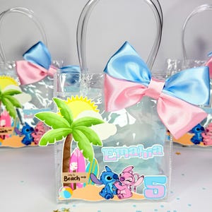 May include: Clear plastic favor bags with blue and pink satin bows. Each bag features a beach-themed design with a palm tree, surfboards, and cartoon characters. The bags have clear handles and the name "Emahra" and the number "5" printed on them.