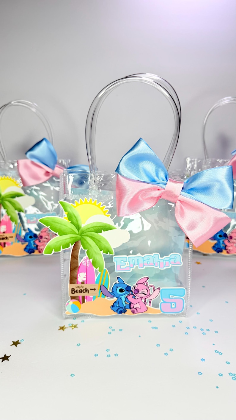 May include: Clear plastic favor bags with blue and pink satin bows. Each bag features a beach-themed design with a palm tree, surfboards, and cartoon characters. The bags have clear handles and the name "Emahra" and the number "5" printed on them.