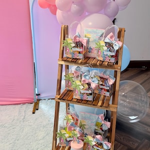 May include: A wooden display shelf holds several gift bags and a small container, all decorated with a cartoon character theme. The bags are clear with colorful bows and palm tree accents. Pink and light blue balloons are in the background.