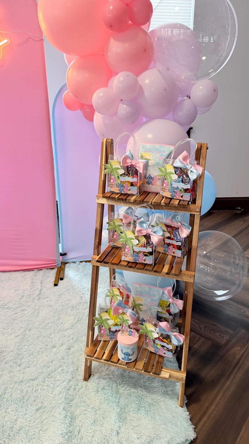 May include: A wooden display shelf holds several gift bags and a small container, all decorated with a cartoon character theme. The bags are clear with colorful bows and palm tree accents. Pink and light blue balloons are in the background.