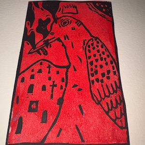 Puede incluir: Un grabado en linóleo rojo y negro que representa un pájaro con una corona y un edificio con cruces. El pájaro tiene un pico largo y patrones de alas detallados. La impresión está sobre un trozo de papel rectangular.