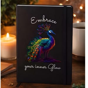 May include: Black journal with a colorful peacock illustration. The peacock has a blue head, a vibrant, multi-colored tail, and the text "Embrace your inner Glow" in white script. The journal has a black elastic band.