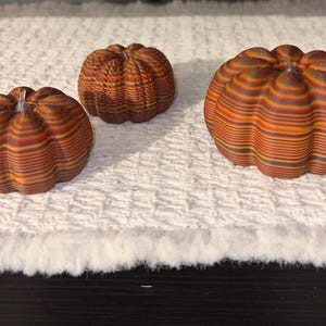 May include: Three decorative pumpkins with a striped design in shades of orange, brown, and green. The pumpkins are arranged on a white, textured surface, perfect for fall decor. The pumpkins vary in size, with the largest one in the foreground.