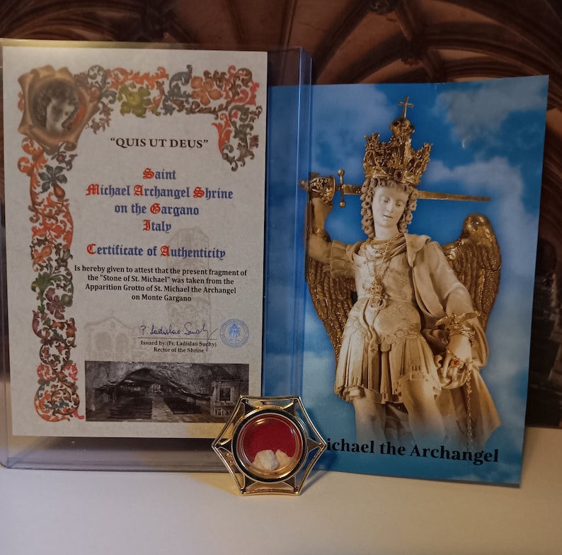 May include: A gold-colored metal reliquary with a red interior and white stone fragments. The reliquary is in front of a certificate of authenticity for a fragment of the "Stone of St. Michael" from the Archangel Michael Shrine on Monte Gargano, Italy. The certificate is signed by the Rector of the Shrine. A picture of the Archangel Michael is in the background.