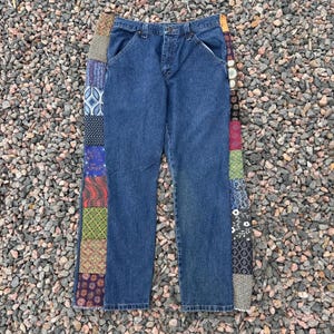 Op de afbeelding: Blauwe spijkerbroek met een patchwork ontwerp aan de zijkant. De jeans hebben een klassiek ontwerp met vijf zakken en een rechte pijp. Het patchwork heeft verschillende patronen en kleuren.