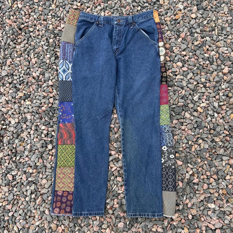 Op de afbeelding: Blauwe spijkerbroek met een patchwork ontwerp aan de zijkant. De jeans hebben een klassiek ontwerp met vijf zakken en een rechte pijp. Het patchwork heeft verschillende patronen en kleuren.