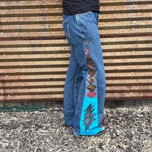 May include: Blue denim jeans with decorative patchwork down the side. The patchwork includes diamond shapes in various colors and a turquoise panel with a black lightning bolt design. The jeans have a classic five-pocket design.