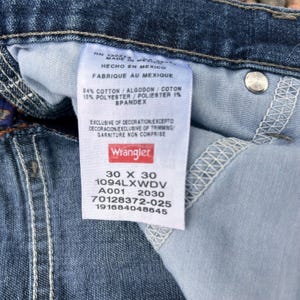 Op de afbeelding: Close-up van een blauw denim jeanslabel met het Wrangler-logo. Het label bevat de maat 30 x 30, de samenstelling van de stof en fabricagedetails. De jeans zijn gemaakt in Mexico.