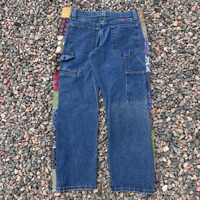 Op de afbeelding: Blauwe denim werkbroek met meerdere zakken en een patchwork ontwerp aan de zijkant. De broek heeft een relaxte pasvorm en een Wrangler label.