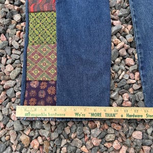 Op de afbeelding: Close-up van een spijkerbroek met een decoratief patchworkpaneel. Het paneel heeft verschillende stoffen met patronen in tinten rood, groen en paars. Een houten liniaal met tekst staat onderaan de afbeelding.