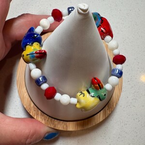 May include: A beaded bracelet featuring small white round beads, faceted red beads, and blue cylindrical beads. The bracelet includes three larger, hand-painted ceramic charms shaped like stylized birds with yellow, blue, green, and red color accents.