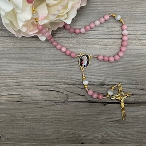 Op de afbeelding: Een roze en witte rozenkrans met een verguld kruisbeeld en een ronde hanger met een religieus beeld. De rozenkrans ligt op een houten oppervlak.